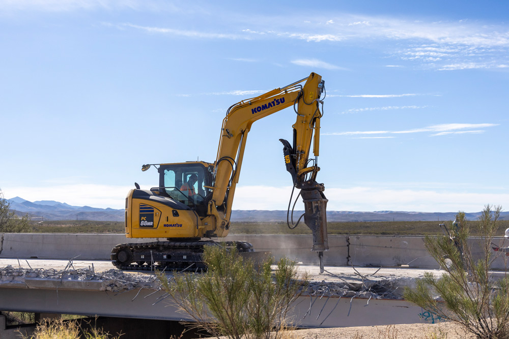 Excavator with a hydraulic breaker demolishing concrete deck on the I-10: Amole TI and UPRR Bridge Rehabilitation Project, with desert landscape and mountains in the background.