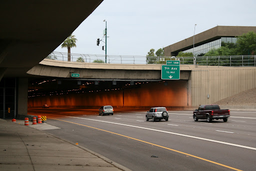Several vehicles drive toward and into a tunnel under an overpass on a multi-lane road. An overhead sign indicates an exit for 7th Avenue.