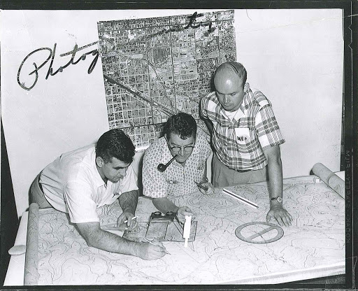 Three men examine a large topographic map on a table, with drafting tools in use; a city map hangs on the wall behind them.