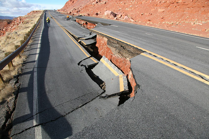 A cracked highway with a large section collapsed, exposing red earth beneath; workers in safety vests are visible in the background near the damaged area.
