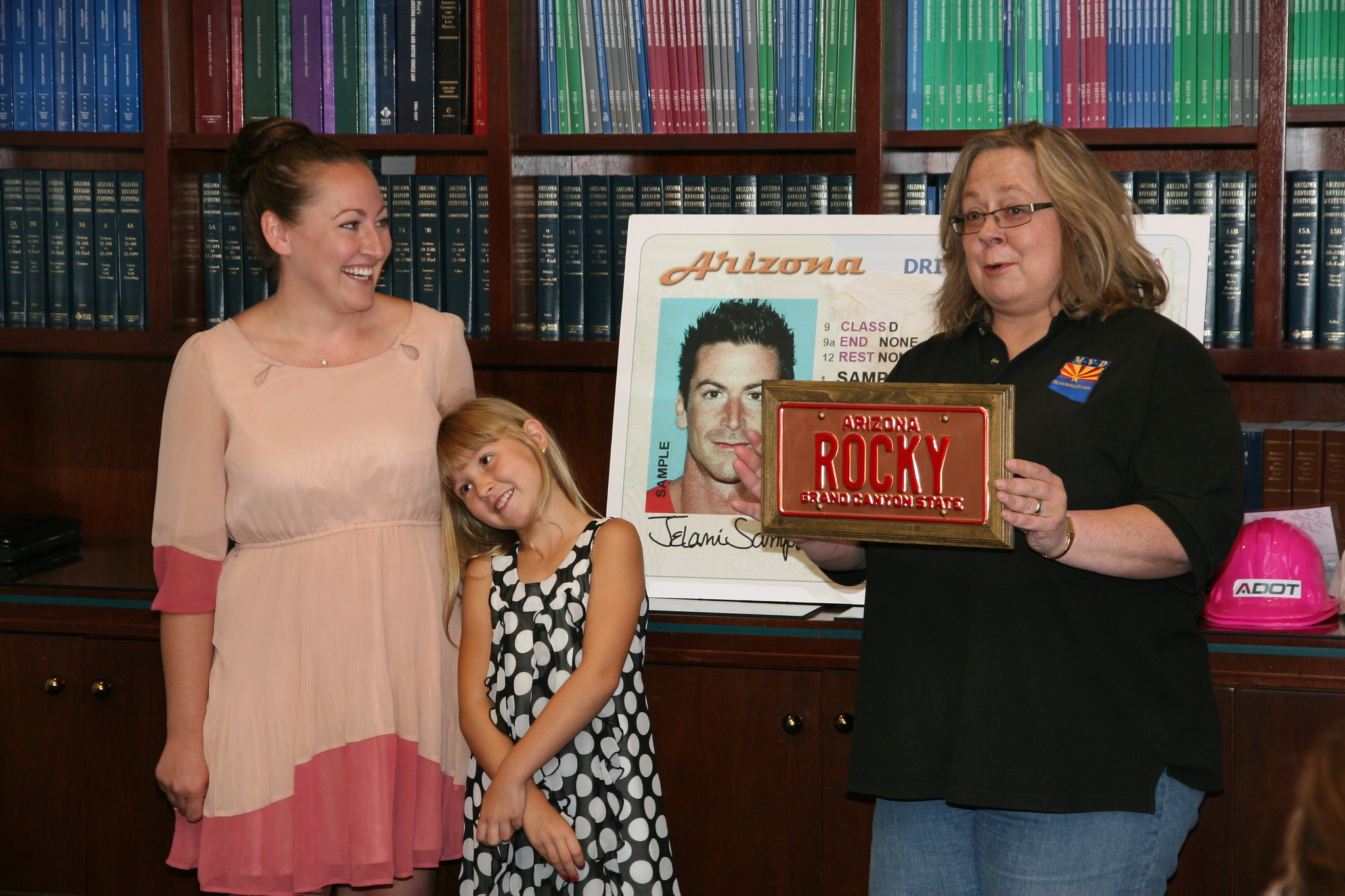 Two women and a young girl stand in front of a large driver license poster and an Arizona license plate.