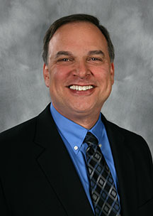 A man in a black suit jacket, blue dress shirt, and patterned tie smiles. He is posed against a gray studio backdrop.