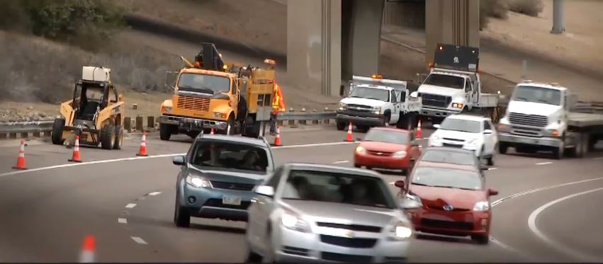 Road construction vehicles and workers on a highway, with traffic cones guiding cars around the work area. Multiple vehicles are moving through the scene.