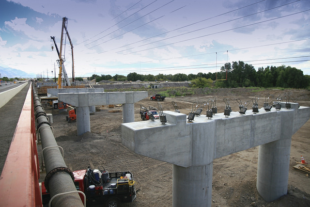 Concrete bridge supports are under construction at a worksite with cranes, vehicles, and equipment; trees and power lines are visible in the background.