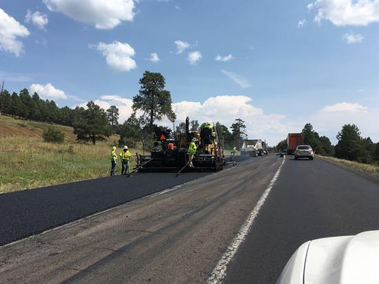 Road construction workers operate paving machinery to lay new asphalt on a highway, with traffic passing by and trees lining the road under a partly cloudy sky.
