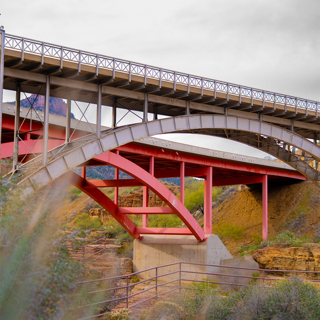 Two steel bridges with arched supports and railings cross over a dry, rocky landscape with sparse vegetation under a cloudy sky.