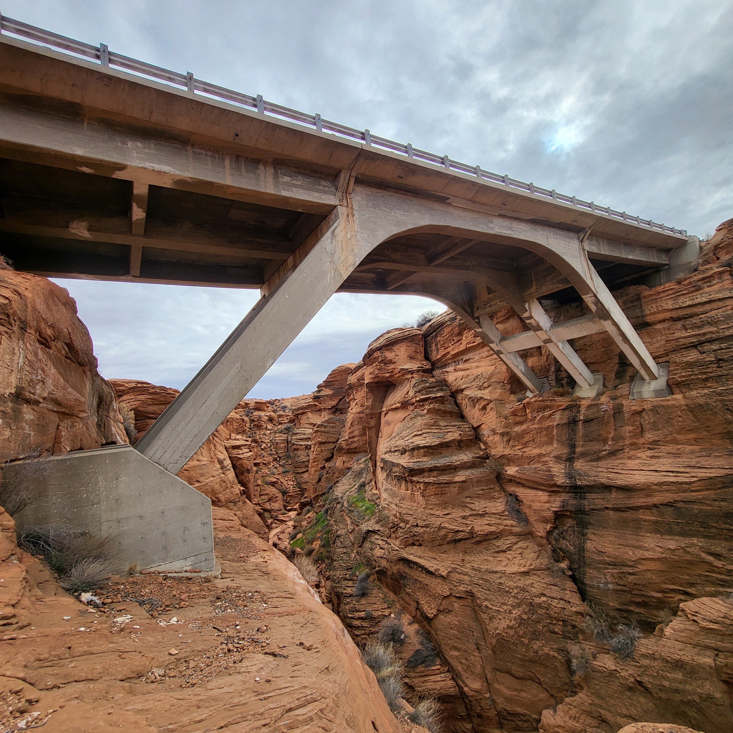 A concrete bridge with large support beams spans a deep red rock canyon under a cloudy sky.