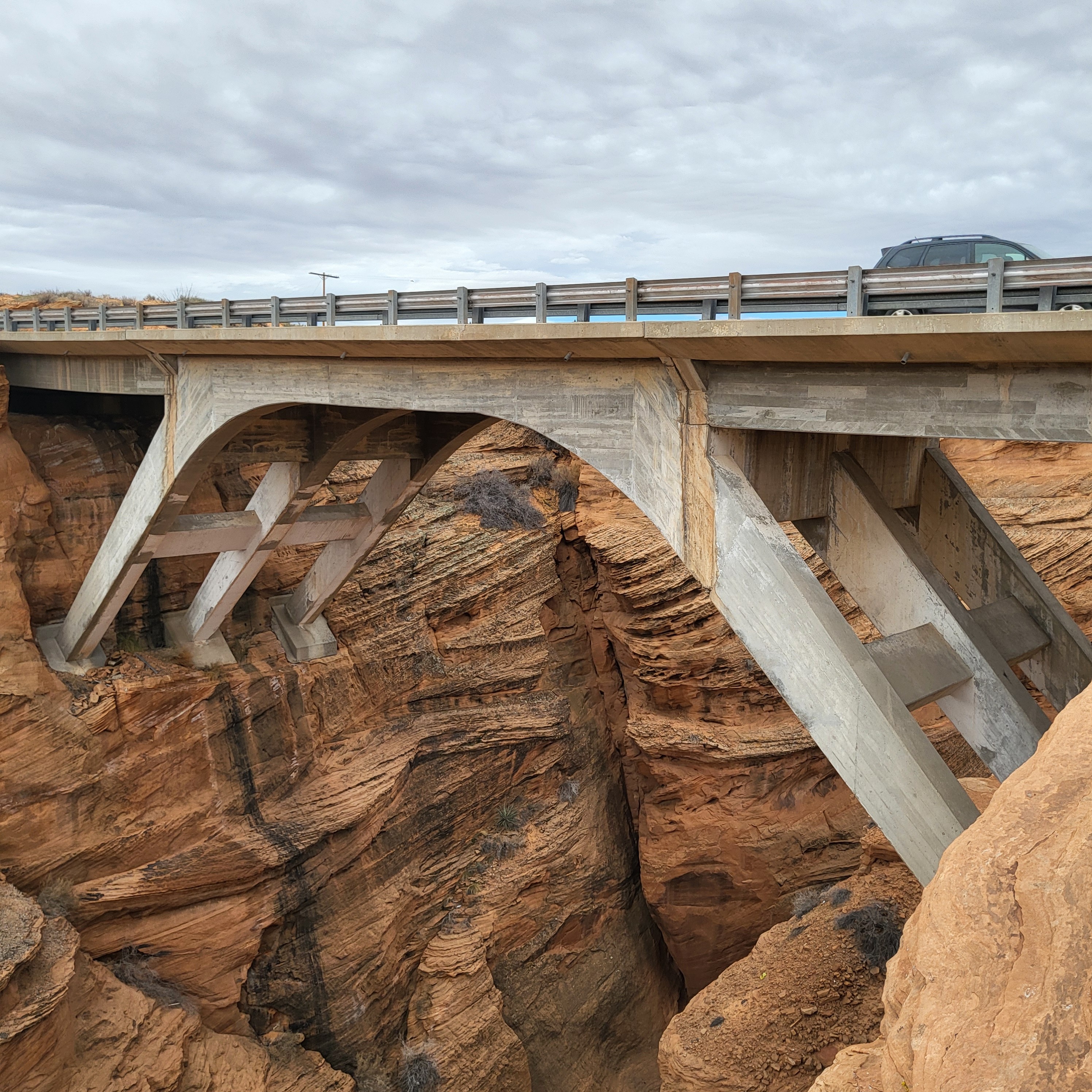 A concrete bridge spans a deep rocky canyon with a car crossing above; cloudy sky overhead and layered rock formations visible below the structure.
