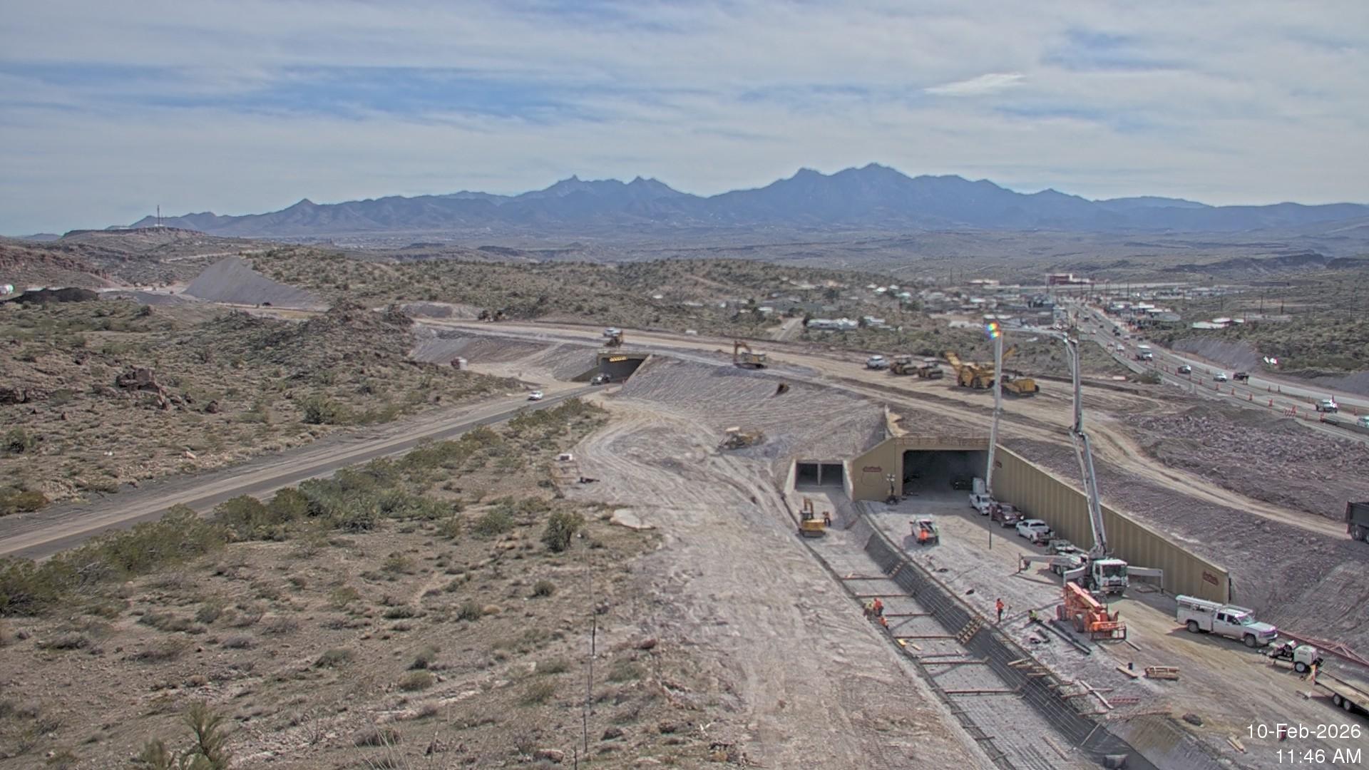 Aerial view of underpass and ramp construction at the I-40 and US 93 West Kingman Traffic Interchange, with heavy equipment, concrete work, and active traffic along the adjacent highway in a desert landscape.