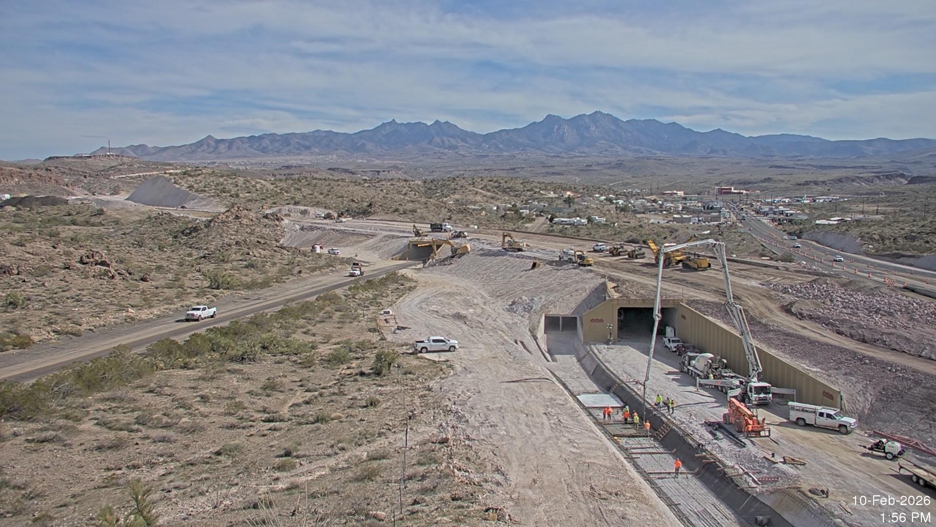 Wide aerial view of underpass construction and ramp earthwork at the I-40 and US 93 West Kingman Traffic Interchange, with concrete pump trucks, heavy equipment, and active highway traffic set against a desert mountain backdrop.