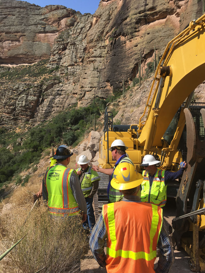excavator operating on a narrow dirt roadway carved into canyon cliffs.