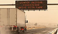 truck driving through a snowy highway with a road sign above saying winter driving conditions drive with care