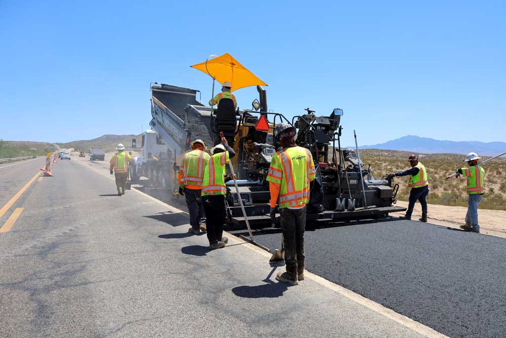 Several road workers lay fresh pavement on US70 using an asphalt paver