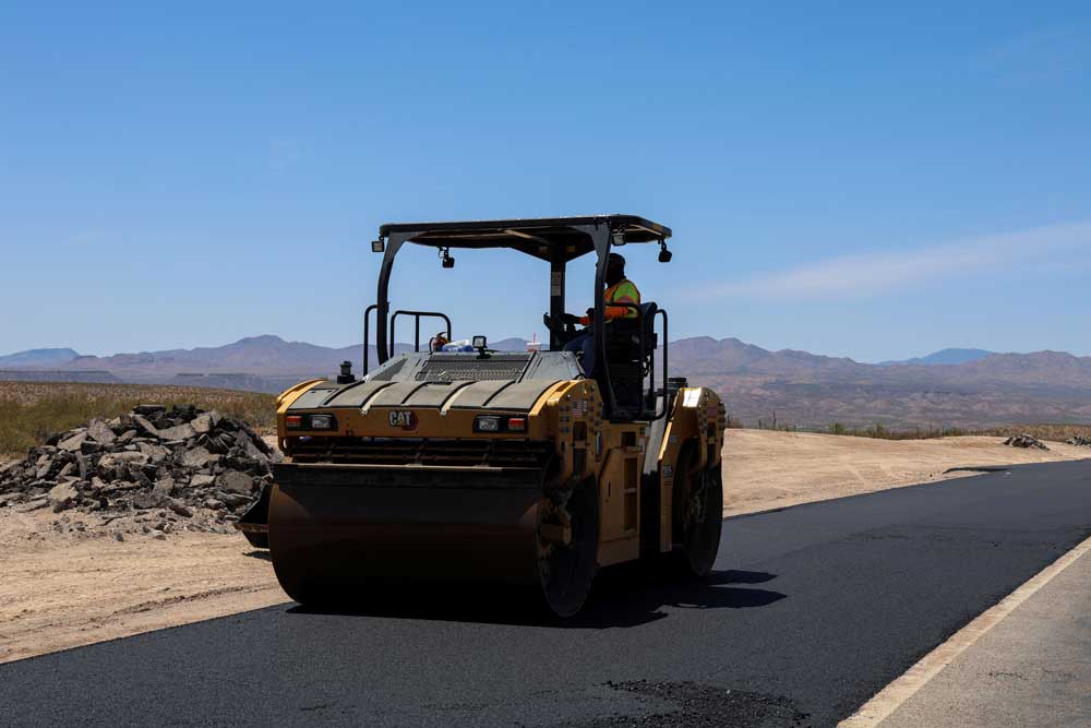 Highway workers operate a compactor roller on fresh pavement on a sunny day.