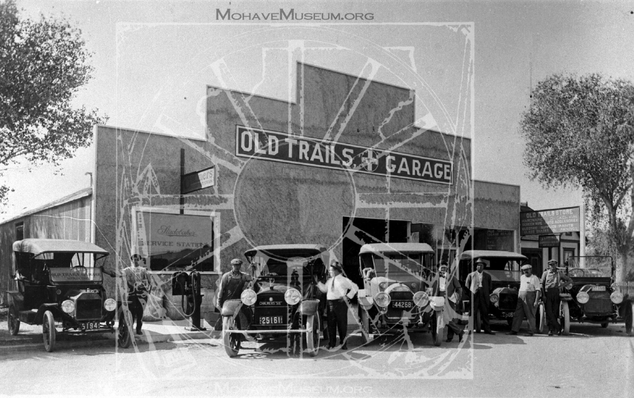 Black and white image of Old Trails Garage. Multiple old vehicles, including Model T's, are parked in front while men stand beside the vehicles.