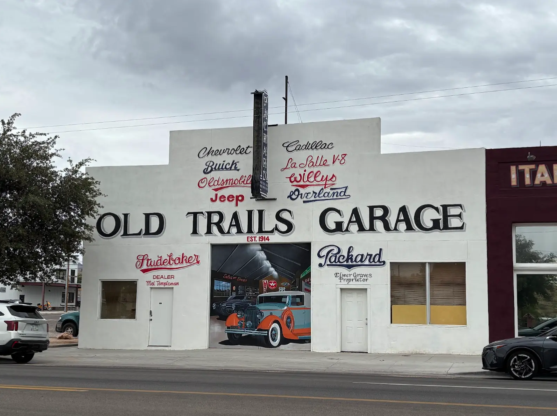 A full color image shows the Old Trails Garage as it is now. The building is white with a mural on the front that appears to look into the garage, with painted vehicles.