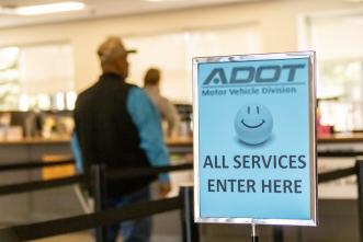 A man stands in line at an Arizona Motor Vehicles Division office. A light blue sign next to the queue features a smiling happy face and the message, "all services enter here."