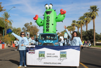 People walk in a parade down a city street. An very tall inflatable cactus can be seen.