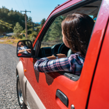 A person in a red car looking out their window to the road in front of them.