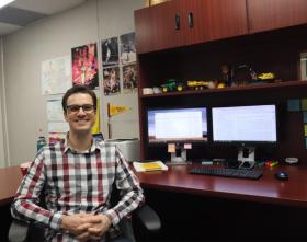 A man sits in a chair in front a desk with computer monitors in an office.