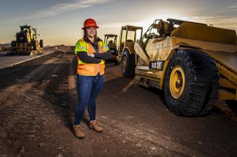 A woman in an orange reflective safety vest stands in a construction area with heavy equipment behind her.