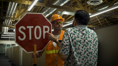 A man in a hard hat and orange vest points at a STOP sign while another man steps back.
