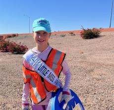 A girl wearing an orange reflective vest poses while picking up litter near a highway.