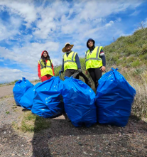 Three people stand behind large, blue trash bags filled with litter removed from a rural highway.