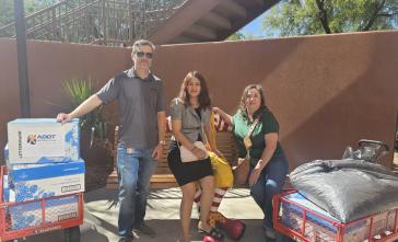 Three people pose for a photo in front of a bench and next to boxes filled with aluminum cab tabs.