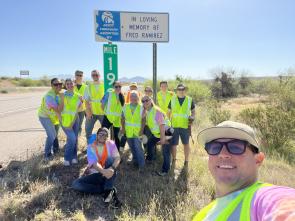 A group of people gather alongside a rural highway, posing for a photo beneath an adopt a highway sign.