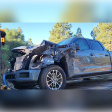 A black truck has severe front end damage. A blue sky and green trees are in the background.