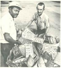Two men stand next two barrels filled with license plates.