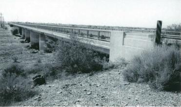 A long, narrow concrete bridge stretches across a dry, rocky landscape with sparse vegetation.