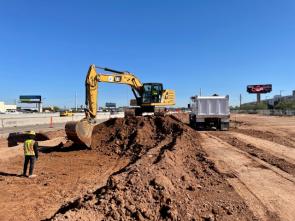 Construction site with an excavator moving dirt into a dump truck. A worker in a reflective vest stands nearby. Clear blue sky in the background.