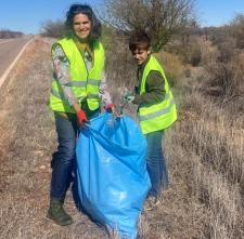 Two people on the shoulder of a rural two-lane highway collect litter and hold a big, blue garbage bag.