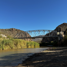 A steel truss bridge spans a calm river with rocky hills and clear blue sky in the background.