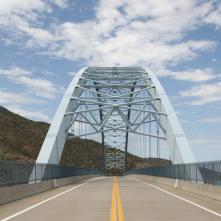 View of a steel arch bridge with a double yellow line road, surrounded by hills and a partly cloudy sky.