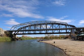 A steel truss bridge spans over a river with a sandy shore and shrubs. A clear blue sky with scattered clouds is in the background.