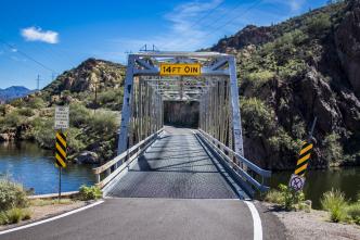 Metal truss bridge over a river, flanked by rocky hills, marked with a clearance sign of "14FT-3IN." Road signs and guardrails are present.