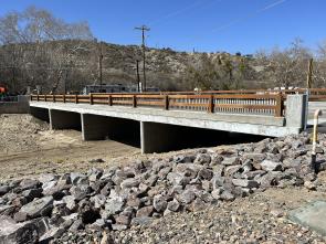 Concrete bridge with brown railings spans a dry, rocky creek bed. Power lines and barren trees are visible in the background under a clear blue sky.