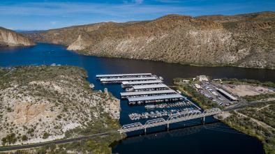 Aerial view of a marina with covered boat docks, a bridge, parking lot, and surrounding desert landscape by a river and rocky hills.