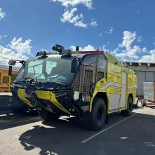 Large yellow airport fire truck labeled "ADOT" parked outdoors on asphalt under a partly cloudy sky, with industrial buildings in the background.