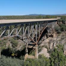 A steel truss bridge spans a rocky canyon with sparse vegetation and distant hills under a clear blue sky.