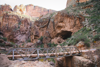 A metal footbridge spans a rocky canyon with steep cliffs and sparse vegetation under a bright sky.