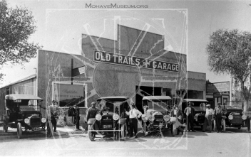 Black and white image of Old Trails Garage. Multiple old vehicles, including Model T's, are parked in front while men stand beside the vehicles.