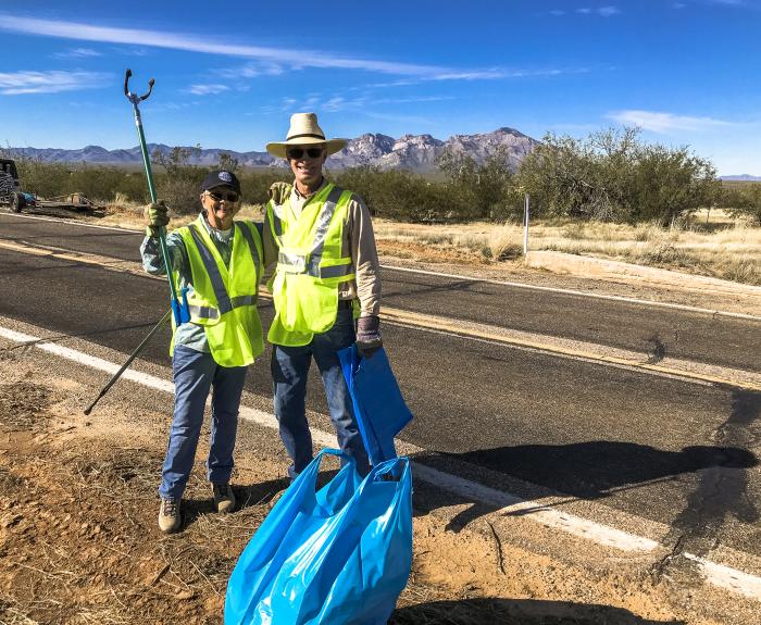 Two people in yellow vests standing on a rural roadside with blue trashbags at their feet.