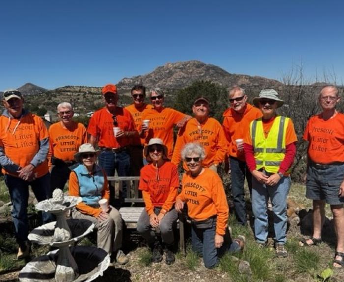 A group of 12 people, all wearing traffic safety orange shirts and members of a volunteer litter-pick-up group, gather for a group photo with a mountain in the background.