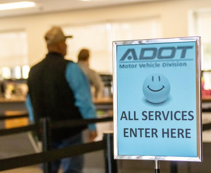 A man stands in line at an Arizona Motor Vehicles Division office. A light blue sign next to the queue features a smiling happy face and the message, "all services enter here."