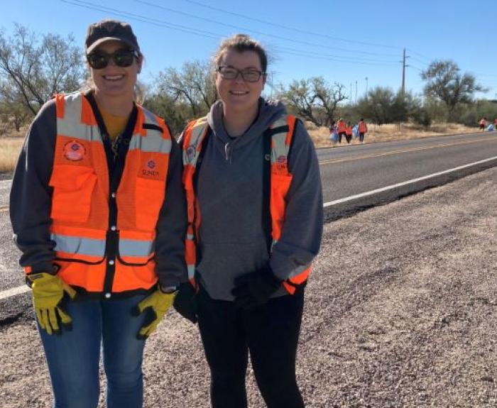 Two people stand on a highway shoulder in orange safety vests. 