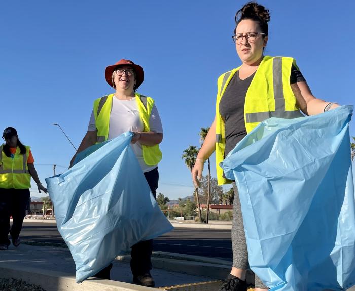 Three people in bright yellow reflective safety vests pick up litter on the side of a highway.